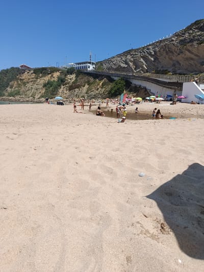 This is a wide, sun-drenched shot of a busy beach in São João das Lampas, Portugal. The scene is dominated by a wide expanse of light-colored sand leading down to the ocean. The sky is a clear, vibrant blue, indicating a sunny day. The time of day appears to be midday or early afternoon, given the bright sunlight and strong shadows.
Numerous people are scattered across the beach, engaging in various activities. Some are sunbathing on the sand, while others are wading or playing in a shallow stream that runs across the beach. There are many colorful beach umbrellas providing shade. In the background, a steep, rocky cliff face rises, topped with lush green vegetation. A modern white building with a prominent antenna sits atop the cliff, overlooking the beach. A wooden boardwalk or walkway winds its way along the side of the cliff, connecting different areas.
The atmosphere is one of leisure and enjoyment, with families and individuals relaxing and having fun on a warm summer day. The presence of the stream suggests a natural water feature meeting the sea. The overall impression is a popular tourist destination with ample natural beauty and amenities. No visible text is present in the image.