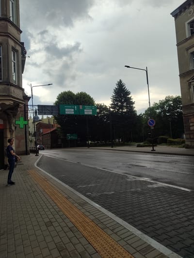 A street scene in Tczew, Poland, features a cloudy sky over a wet asphalt road and a brick sidewalk. A person stands on the sidewalk, looking away from the camera. Buildings line the street, including a structure with a green cross symbol. Road signs indicate directions to various locations, such as "Nowy Dwór Gd", "Centrum", "Oś. Staszica", "Bydgoszcz", "Malbork", "Częstochowa", and "Górki". A blue and red circular traffic sign with a diagonal line is also visible. Trees and streetlights are present along the roadside.