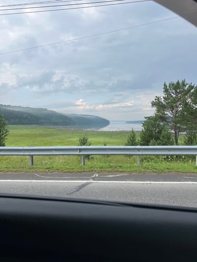 The image depicts a scenic view of a body of water, likely a river or lake, bordered by lush green fields and dense forests. The water is calm and reflects the overcast sky, which is filled with soft, diffused clouds. A winding road with a silver guardrail runs horizontally across the foreground, separating the road from the natural landscape. The vegetation appears vibrant and healthy, with various shades of green in the grass and trees. The overall atmosphere is serene and tranquil, suggesting a rural or natural setting. This scene is located in Patrony, Russia, a place known for its natural beauty and proximity to water bodies.