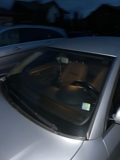 The image depicts an outdoor scene at night in Madzhare, Bulgaria, showing two parked cars. In the foreground, a silver-grey sedan is viewed from a high angle, focusing on its front-right quarter. The car's windshield reveals a dark interior with a tan-colored passenger seat and a black steering wheel partially visible. An object hangs from the rearview mirror. A rectangular holographic sticker, displaying various colors, is affixed to the lower right corner of the windshield. The car's hood, A-pillar, and part of the front fender are discernible. To the left and slightly behind the primary vehicle, another silver-grey car is partially visible, showing its side profile including a door handle and window line. The background consists of heavily blurred, dark shapes that appear to be houses or buildings under a dark sky, indicating low ambient light conditions. A faint red light is present in the distant background. No individuals are visible in the scene.