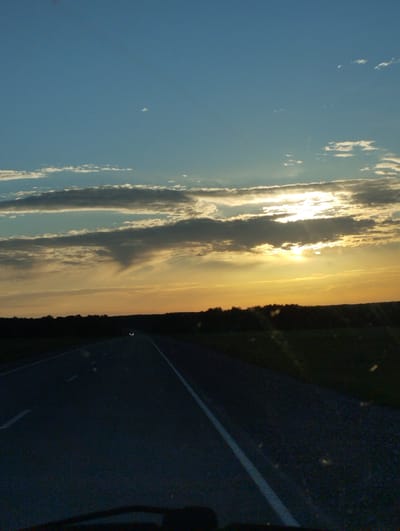 The image depicts a view from inside a vehicle, likely a car, driving on a paved road towards a horizon. The road is marked with white lines. The sky is mostly blue with white and gray clouds. The sun is near the horizon, creating a sunset effect with a yellow-orange glow. A dark, indistinct treeline or vegetation appears in the middle distance. The specific location is not clearly discernible as Filippovo, Russia without more distinct visual features.