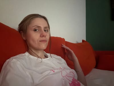 A young woman with light brown hair is lying on a couch with bright orange pillows. She is wearing a white t-shirt with a pink floral design. Her hair is damp and pulled back, and she has a light expression on her face. The background is a wall that is half white and half dark green. The scene appears to be indoors, possibly in a home. There is no indication of the specific location within the city of Helmond, Netherlands, or any environmental context beyond the immediate surroundings.