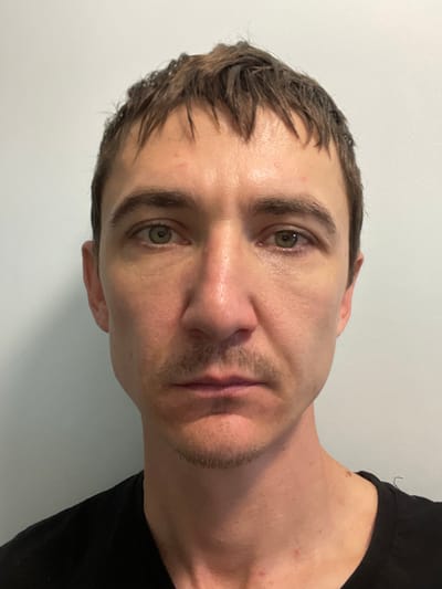 The image is a frontal, medium shot of a man's face, from the shoulders up. He has light skin, short, dark, messy hair, and green eyes. He is wearing a black t-shirt. The background is a plain, light-colored wall. The lighting is even, with no harsh shadows. The image is in focus and shows clear detail of the man's features. There is no visible location information in the image to suggest it is in Laishevo, Russia.
