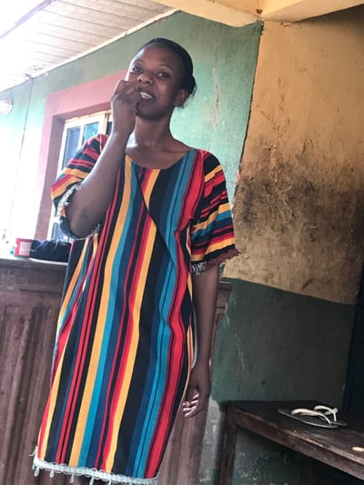 A woman wearing a colorful striped dress stands indoors with a finger near her mouth, appearing thoughtful. The dress features horizontal stripes in blue, yellow, red, and black, with decorative trim on the sleeves and hem. Behind her, a window and wooden door are visible, along with walls painted in green and yellow. A wooden cabinet with white handles can also be seen in the background.