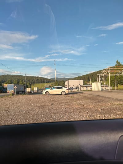 The image depicts a bright, sunny day in Inzer, Russia, with a clear blue sky dotted with white clouds. The foreground is a gravel lot, and a white car is parked in the middle of the frame. In the background, a long, light-colored truck is parked on the side of the road. A forest-covered hill forms the backdrop of the scene, with some buildings and utility poles visible. The overall impression is one of a rural or semi-urban area, possibly a truck stop or rest area.