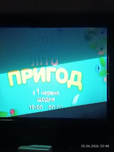 The image shows a television screen displaying a promotional graphic for a show titled "Літо Пригод" (Summer of Adventures). The graphic features large, colorful, stylized text in yellow and orange against a bright cyan background. The text "Літо" is positioned above "ПРИГОД". Below the title, smaller white text reads "з 1 червня щодня" (from June 1st daily) and "19:00 - 00:00" indicating the show's premiere date and daily broadcast times. In the top left corner, a logo resembling a stylized "K1" is visible, and in the top right corner, the letters "HTH" are present. The background is decorated with abstract shapes and colors, suggesting a playful and summery theme. The overall impression is a bright and inviting advertisement for a television program. The timestamp at the bottom right corner indicates the photo was taken on June 25, 2026, at 23:48. The image itself is a photograph of a television screen, likely taken in a dimly lit room, as evidenced by the darkness surrounding the screen and the visible reflection of the environment on the screen. The location context is Bolhrad, Ukraine.