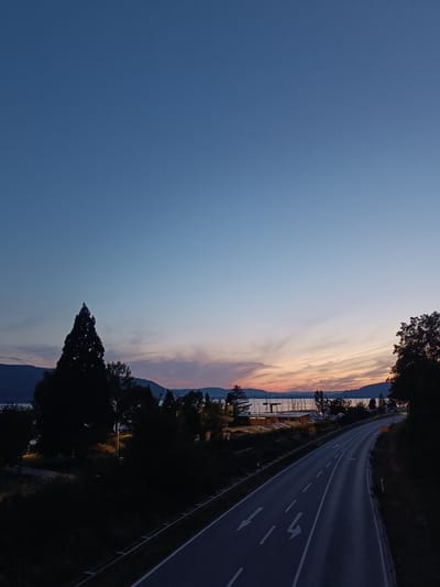 The image depicts a scenic view in Sipplingen, Germany, likely taken at dusk. A road curves through the foreground, with white lane markings indicating a two-lane highway. The road is flanked by dark vegetation and trees on the right, and a more open area with trees and a large, dark conifer on the left. In the middle ground, a body of water, possibly Lake Constance, is visible, with mountains or hills forming a distant shoreline. A harbor with numerous sailboats and a building complex with lights is situated along the water's edge. The sky above is a gradient of deep blue at the top, transitioning to lighter blues and then displaying subtle hues of orange and pink from the setting sun. There are no people or animals visible in the image.