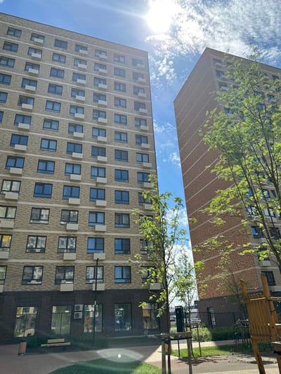 This is a medium-angle, eye-level shot of a residential complex in Znamya Oktyabrya, Russia, on a sunny day. The scene is dominated by two multi-story buildings. The building on the left is a tall apartment block with a light-colored brick facade, featuring numerous windows and air conditioning units. The building on the right is a similarly tall apartment block with a reddish-brown brick facade, also adorned with windows and air conditioning units.

In the foreground, a paved area transitions into a green lawn with scattered trees. To the right, a wooden playground structure is visible, partially obscured by foliage. A black trash bin and a wooden bench are situated near the base of the left building, adjacent to a walkway. The sky above is a bright blue, with scattered white clouds and the sun prominently visible, creating lens flare. The overall impression is one of a modern urban residential area bathed in sunlight.