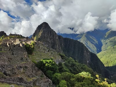 Machu Picchu stands majestically amidst the Andes mountains. The ancient Inca citadel is terraced, with stone structures cascading down the slope. Lush green vegetation contrasts with the grey stonework. A towering mountain peak looms over the site, partially obscured by swirling clouds. In the distance, other verdant peaks and valleys stretch into the horizon, creating a stunning landscape under a cloudy sky.