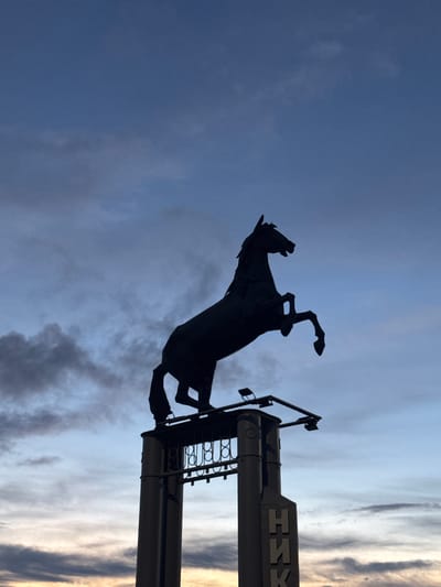The image depicts a silhouette of a horse sculpture against a dusky sky. The horse is rearing up, its front legs extended and its head held high, conveying a sense of power and dynamism. It is positioned in front of two large, cylindrical structures, likely pylons or columns, which are topped with decorative elements and lighting fixtures.
On the rightmost pylon, a large, stylized emblem is visible. The emblem consists of geometric shapes forming what appears to be letters. While the full text is not discernible, the shapes suggest a possible interpretation of "NKH" or similar characters, possibly indicating a name or affiliation. The overall scene is illuminated by soft, diffused light, suggesting it is either dawn or dusk. The sky is a mix of deep blue and lighter, cloud-streaked areas, with hints of warmer tones near the horizon, further reinforcing the time of day. The image is shot from a low angle, emphasizing the scale of the sculpture and the supporting structures against the vastness of the sky. The setting appears to be an outdoor public space, given the monumental nature of the sculpture and its placement.