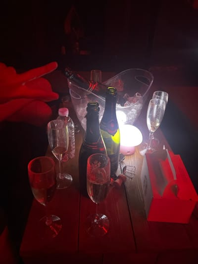 The image displays an indoor scene within a dimly lit establishment, likely a lounge or club, located in 강남구, South Korea. The environment is dominated by a red ambient light, illuminating a wooden table surface where several items are arranged.
Central to the scene is a clear plastic ice bucket containing ice and one dark-colored bottle, possibly champagne or sparkling wine, partially submerged. Adjacent to the bucket, two additional dark glass bottles of champagne or sparkling wine stand upright; one appears uncorked, with its cork and wire cage resting on the table nearby. A clear plastic water bottle with a pink cap is also visible.
Five champagne flutes are present on the table; two contain a significant volume of light-colored liquid, consistent with champagne, while the remaining three appear to be partially consumed or empty, showing residual liquid. A small, clear drinking glass is also observed near the ice bucket. A compact, mushroom-shaped portable lamp, emitting a soft white-pink glow, is positioned amongst the bottles. On the right side of the table, a red and white cardboard box is situated.
A person's left hand and forearm, cast in red light, are visible on the far left, with the index finger extended and pointing towards the upper center of the frame. No other individuals are discernibly present.