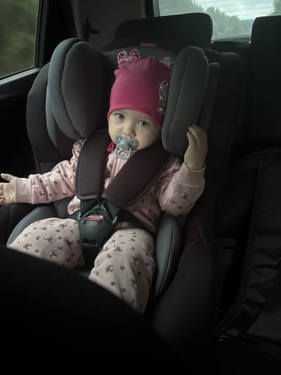 A baby wearing a pink hat and pajamas with a pattern, secured in a gray car seat with a dark gray harness, is visible inside a vehicle. The baby has a pacifier in its mouth. The location is presumably Михайловка, Russia. A car window with trees visible outside is in the background. A dark object, possibly a bag or seat, is above the baby.