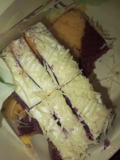 A box of sliced cake features an eye-catching color contrast. The cake has alternating layers of yellow and purple sponge. A generous layer of white frosting covers the cake, adorned with shredded white cheese. The slices are neatly arranged in the box, with a stray piece lying separately. The image is taken from an overhead angle, providing a close-up view of the sweet treat.