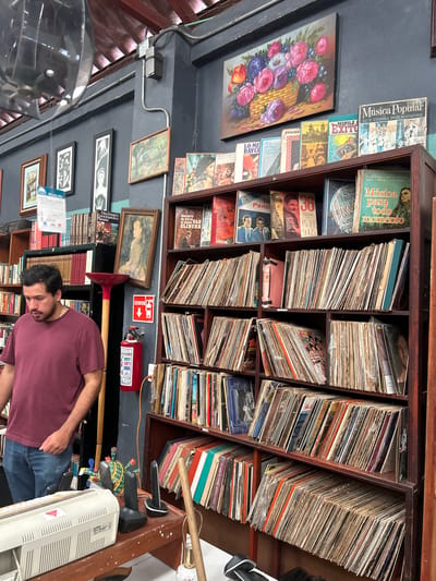 Here's a detailed description of the image:
**Setting:** The image appears to be taken inside a used bookstore or antique shop, potentially located in Veracruz City, Mexico, based on signage written in Spanish and general aesthetic.
**Composition:** The shot is an interior view with a slightly rotated perspective. A man is situated in the lower center of the frame. The primary focus is a tall, packed bookshelf filling the upper portion of the image.
**Bookcase Contents:** The bookshelf is loaded with old books, vinyl records, and magazines. Some items have Spanish text on their covers, suggesting they are related to Mexican culture. Titles include 'Música Popular,' 'EXITO,' and 'Diccionario Enciclopédico Planeta.' The items are tightly packed and appear to be used or aged.
**Man:** A man with dark hair and a beard is in the lower part of the frame, wearing a maroon shirt and jeans. He appears to be browsing the books or looking for something. His face is slightly turned downward.
**Other Objects:** A red fire extinguisher and emergency exit sign are affixed to the wall near the bookshelf. There are also some framed paintings hanging on the wall, adding to the vintage and eclectic atmosphere of the store. Also near the bottom right of the image is a vintage computer.
**Lighting:** The lighting is diffused, suggesting indoor lighting within the shop. The light is not particularly bright.