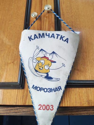 A white pennant with blue and white trim hangs from a wooden surface, likely a door. The pennant features a cartoon image of a skier with a yellow face, red hat, and blue skis, against a backdrop of mountains and a skier's trail. Above the image, the word "КАМЧАТКА" (Kamchatka) is printed in blue capital letters. Below the image, the word "МОРОЗНАЯ" (MoroZnaya - Frosty) is also in blue. At the bottom of the pennant, the year "2003" is printed in red. The wooden surface has a visible wood grain pattern and two silver fasteners holding the pennant's cord. The background appears to be an interior setting, possibly a home or a lodge, with a black frame visible to the left of the pennant. The overall impression is of a souvenir or decoration related to a winter sport or travel to the Kamchatka region of Russia.