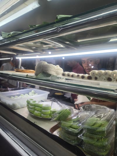 This is a photograph taken inside a food display case, likely in a market or restaurant in the city of Medan, Indonesia. The display case has multiple shelves, illuminated by fluorescent lights. On the top shelf, a pile of white, stringy food, possibly a type of noodle or sweet confection, sits on a green leaf. Next to it, a row of small, white, cylindrical pastries with dark centers are arranged on a white plate.
On the shelf below, a white plastic container is filled with small, pale green, ball-shaped sweets dusted with white powder. In front of this container, a stack of green, ring-shaped cakes, also dusted with white, are arranged on a white plate. To the right of these cakes, several clear plastic containers are stacked, each filled with green, flat, disc-shaped pastries.
In the background, two individuals are visible. One, wearing a white shirt with orange stripes, appears to be standing behind the display. Another, a young man with dark hair, is looking directly at the camera through the glass of the display case. The overall environment suggests a commercial food vendor.