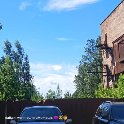 An outdoor, eye-level, medium shot shows the top of a silver car in the foreground. A dark brown wooden fence runs horizontally across the middle of the frame, behind which are trees with lush green foliage and a tall, light brown brick building with a dark metal fire escape. The sky is a clear, bright blue with scattered white clouds. Text in white Russian characters, "найди меня если сможешь" (find me if you can), appears on the car's windshield, accompanied by three emoji: a purple devil, a yellow face with sunglasses, and a brown cowboy. The car is parked in what appears to be a parking area or a yard. This scene is not specific to Pskov, Russia, and could be anywhere.