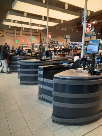 The image shows a row of checkout counters in a supermarket in Rosenheim, Germany. The supermarket appears to be a Lidl, as indicated by the logo visible on a sign in the background that reads "Vielen Dank für deine Einkauf." Several people are present, including shoppers and possibly supermarket employees. One person in the foreground is wearing a white cap and grey sweatpants, standing near a checkout counter. Another person, wearing a green t-shirt, is visible behind a counter, likely working as a cashier. The checkout counters are dark blue with grey trim. A computer monitor, payment terminal, and scanner are visible on one of the counters. The environment is brightly lit by overhead fluorescent lights. The floor is tiled in a light beige color. Several signs are visible in the background, some with German text. The overall scene depicts a typical supermarket checkout area in operation.