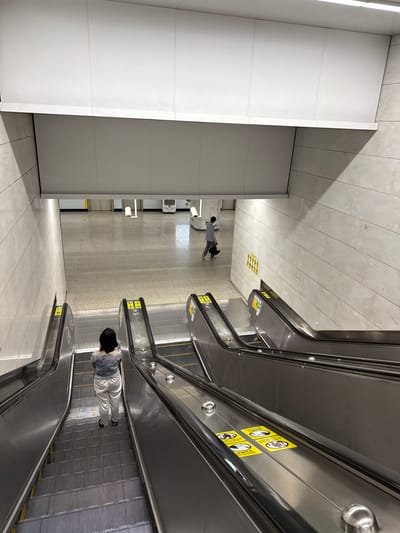 In a subway station in Liwan District, China, a woman with dark hair stands on a moving escalator. She is wearing a light blue and white patterned shirt and white pants. She is facing away from the camera, looking down the escalator. In the background, a man walks across the station floor, carrying a black bag. The station has a polished stone floor and white tiled walls. Several other escalators are visible, some with yellow safety signs.