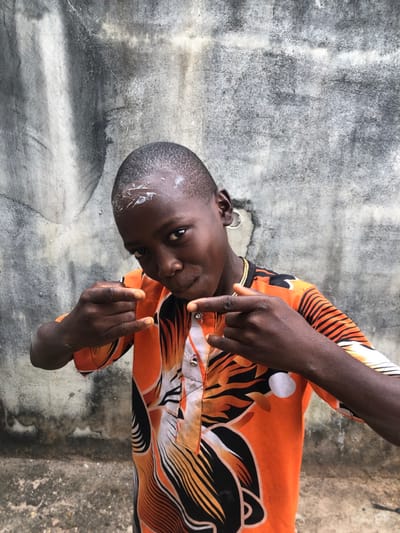 This image, captured in Oraukwu, Nigeria, presents a young male subject standing centered against a weathered concrete wall. The individual has dark skin, a short buzz cut, and a white substance visible on his forehead. He wears a thin gold-colored chain around his neck and a silver hoop earring in his visible right ear. His attire includes a short-sleeved, collared shirt featuring an orange, black, and white abstract or animal-print pattern, with a v-neck closure secured by two silver buttons. The subject's hands are raised to chest level, each forming a 'horns' gesture with extended index and pinky fingers. He maintains direct eye contact with the camera, exhibiting a slight smirk. The background wall is light gray, exhibiting extensive dark streaking, staining, and textural inconsistencies.