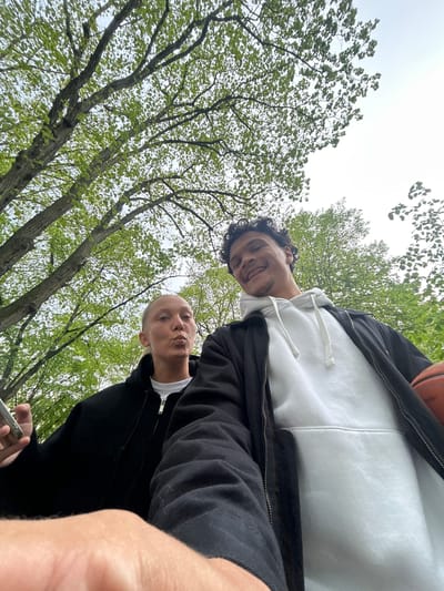 Two people are looking down at the camera from a low-angle perspective, likely in an outdoor park setting. The person at the top of the frame is smiling and wearing a white hooded sweatshirt with a black zip-up jacket. They are holding a basketball partially visible in the top right corner. The person below is making a kissing face and wearing a white t-shirt with a black zip-up jacket. They are holding a smartphone in their hand, presumably taking the picture. The background consists of trees with green foliage. The photo was taken during the day, under an overcast sky. It's difficult to be certain of the location in Noisiel, France, but the style of the trees suggests it could be a park.