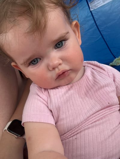 A close-up, waist-up shot focuses on a baby with light brown hair and blue eyes, wearing a pink ribbed onesie. The baby is looking directly at the camera with a neutral expression. The background is blurred, with a large section of blue fabric and a hint of green. The lighting is natural and suggests an outdoor setting. There are no discernible Giessen landmarks or any contextual clues to indicate the location. The image is a portrait of the baby.