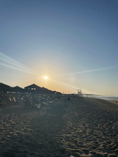 The image shows a beach scene at sunset in an unknown location in Greece. The sun is low in the sky, casting a warm glow over the scene. In the foreground, there are rows of empty beach chairs and umbrellas set up on the sand. Further down the beach, a lifeguard tower stands tall. The sea is calm, and there are no people visible in the water. The sand is textured with ripples and shadows. The overall impression is one of tranquility and relaxation.