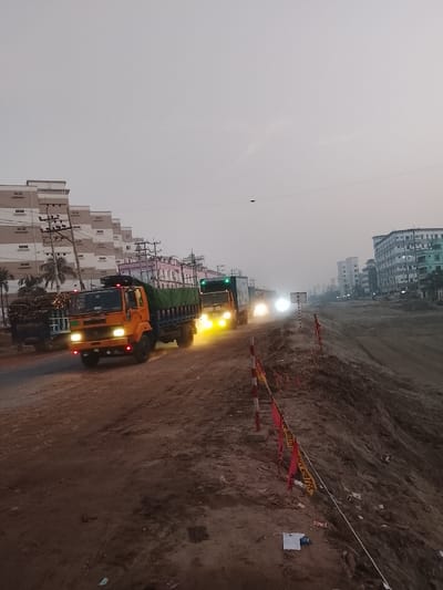 A line of trucks drives along a dirt road during twilight. The lead truck is orange and is followed by other trucks with covered beds. The road is bordered by a dirt bank with warning flags. Buildings line the left side of the road and more distant buildings are on the right. The sky is overcast.