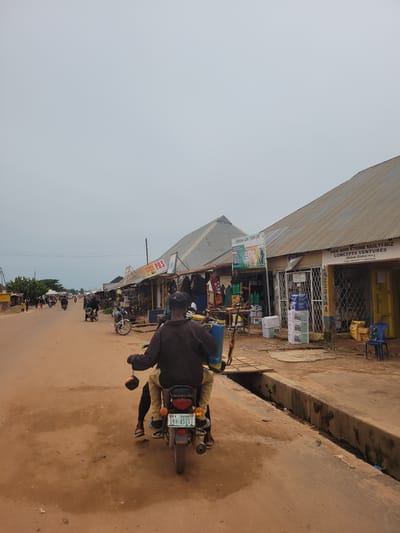 This is a sideways, eye-level, full-shot photograph taken in Bokkos, Nigeria. The scene depicts a roadside marketplace with several shops lining the street. The shops have corrugated metal roofs and are filled with various goods, including what appear to be agricultural supplies and electronic items.
A man is the primary subject, seen from behind riding a motorcycle down the dirt road. He is wearing a dark jacket and light-colored pants. The motorcycle has a license plate that reads "BKK-4510T" and "PLATEAU". Other motorcycles and a few people are visible further down the street, suggesting a busy day.
The sky is overcast and light, indicating it might be morning or late afternoon, and the weather appears to be neutral. The road is dusty and unpaved, typical of many areas in Nigeria. The overall atmosphere is one of everyday commerce and activity in a local market setting.
Visible text includes shop names such as "OAK AND STONE MULTI-BIZ CONCEPTS VENTURES," "CENTEENO AGRO TECH LTD," and signs indicating "RECHARGE CARD SALES" and "P.O.S." There are also contact phone numbers displayed on one of the shop signs. The image captures a slice of daily life in this Nigerian town.