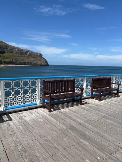 The image depicts a pier scene in Llandudno, United Kingdom, on a sunny day. Two dark brown wooden benches with backrests are positioned along the edge of the pier, separated by a patterned white and blue railing. The pier itself is constructed of light-colored wooden planks, with visible nails. Beyond the railing is the sea, with a coastline featuring a rocky, green-topped cliff face. The sky is blue with scattered white clouds. There are no people visible in the shot.