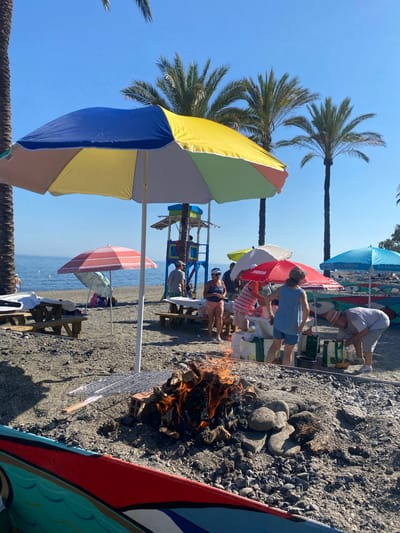 The image depicts a lively beach scene in Marbella, Spain, on a sunny day. A group of people are gathered on a pebbly beach, enjoying a barbecue. In the foreground, a fire pit with burning wood and stones is central to the activity, with a metal grill positioned above it. Several colorful beach umbrellas, including a large multi-colored one and a red one, provide shade for the attendees.
People are seen interacting around a table laden with food items and shopping bags. One individual is tending to the fire, while others are either preparing food or observing. In the background, palm trees line the shore, and the Mediterranean Sea stretches out under a clear blue sky. A lifeguard tower is visible further down the beach, suggesting the area is a popular recreational spot. The overall atmosphere is one of casual outdoor gathering and enjoyment.