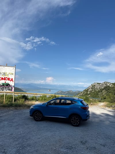 A bright blue car is parked on a gravel road overlooking a vast, mountainous landscape with a body of water in the distance. The car is positioned on the right side of the frame, facing away from the viewer. Its wheels and part of its side profile are visible.
The scene is set during the daytime, with a clear blue sky dotted with white clouds. The sun is shining, casting light on the car and the surrounding environment. To the left of the car, there is a steep, rocky mountain covered in greenery. In the background, a hazy, expansive view of mountains and a large body of water unfolds, suggesting a coastal or lake region.
In the foreground, on the lower left side of the image, a sign with graffiti is visible. Parts of the text on the sign are legible, including "IT'S TIME FOR" and "ONDRA," along with some abstract graffiti art in bright colors. A yellow barrier fence runs along the edge of the road, separating it from the drop-off with the view. The overall impression is one of a scenic overlook, possibly a viewpoint or a stop along a mountain road. The setting appears serene and offers a panoramic vista.