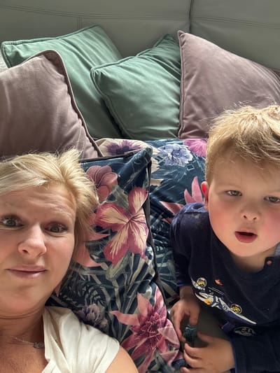 A selfie captures a woman with short blonde hair, lying on a floral-patterned pillow alongside a young child. The child, with blonde hair and fair skin, is wearing a navy blue pajama top with a rocket ship design and holding a green dinosaur toy. They are positioned on top of the floral pillow, while the woman is positioned below. The floral pillow is a mix of dark blue, green, and pink flowers. The background includes a stack of plush pillows in lavender and teal. The location is not identified, but the person taking the photo seems to be indoors and the environment appears to be a home interior.