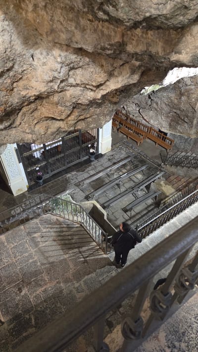 A high-angle shot depicts the interior of a structure in Altura, Spain, possibly a cave or a deep well, with rough-hewn rock walls on the left. A stone stairway with metal railings zigzags down into the depths of the structure. A person in dark clothing and shoes stands on a landing with a metal railing, looking down. The overall perspective is disorienting, as the image is tilted. A wooden structure is visible at one level of the descent. A lower level has a white structure with windows and potted plants.