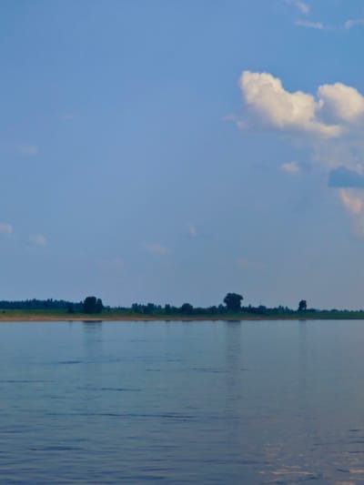 The image depicts a wide, calm river under a partly cloudy sky. A thin strip of land with a dense line of trees forms the horizon. The sky is a gradient of blue, lighter towards the horizon and deeper overhead, with a few fluffy white clouds scattered across it. The water is a muted blue-grey, reflecting the sky and showing subtle ripples. The overall impression is one of natural tranquility. The location is identified as Erkalovo, Russia, suggesting a riverside scene in that region. There are no people or visible signs of human activity within the frame.