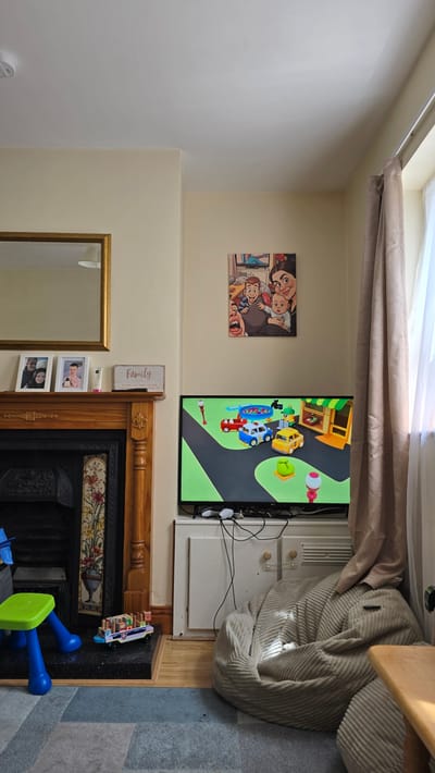 The image depicts an interior living room scene, likely a home, in Kilkenny, Ireland. The room is furnished with a large television displaying a cartoon, a fireplace with a mantelpiece adorned with picture frames and a decorative sign, a beanbag chair, and children's toys. A window with curtains is visible on the right side of the frame. The overall ambiance suggests a relaxed, family-oriented environment.