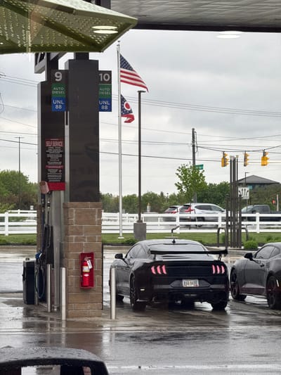 This media file captures an outdoor scene at a gas station under overcast conditions, with a wet ground suggesting recent rain. The time of day appears to be daytime.
In the foreground, a black sports car, possibly a Ford Mustang, is prominently featured from the rear, displaying a license plate partially reading "KVB 9456". It has a spoiler and distinctive red LED taillights. Another dark grey car is parked further ahead.
To the left, two gas pumps, numbered "9" and "10," are visible, displaying "AUTO DIESEL" and "UNL 88" (unleaded 88 octane) prices. Above these, an American flag and the distinctive Ohio state flag are flying, indicating the location is in Ohio, USA. A prominent "SELF SERVICE" sign provides various warnings, including "NO SMOKING STOP ENGINE," cautions about dispensing gasoline into unapproved containers, and warnings about static electricity.
The background shows traffic lights with yellow casings, power lines, and white picket fences lining the roadside next to green foliage. A red fire extinguisher is mounted on a brick pillar near the pumps. The scene depicts a functional gas station setting during inclement weather.