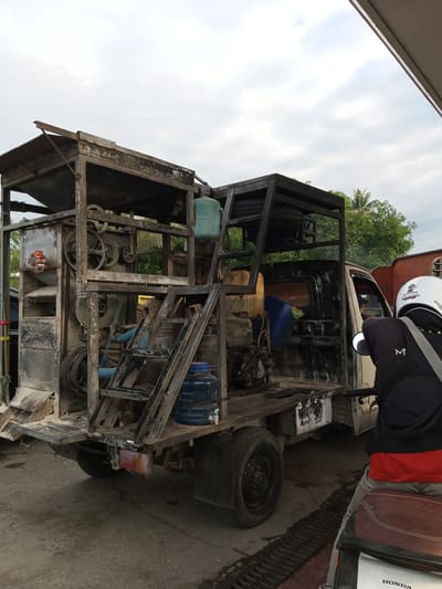 In the city of Tocina, Indonesia, a modified pickup truck is parked. The truck bed is heavily customized with a large, elaborate metal frame structure, appearing to house some kind of processing or mechanical equipment. Various tanks, pipes, and machinery components are visible within this structure. The truck itself is weathered and shows signs of extensive use. In the foreground, a person is seated on a motorcycle, facing away from the camera. The person is wearing a helmet and dark clothing. The environment appears to be an outdoor area, possibly a service station or workshop, with a concrete ground and a covered overhang. Trees are visible in the background. The sky is overcast with clouds.