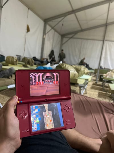 A person is sitting on a cot in a large white tent, playing a video game on a red Nintendo DS handheld console. The game is showing a racing track with two vehicles driving on it. The person is holding the console with their right hand. The upper screen of the console is displaying the racing track, while the lower screen displays a game map with a list of numbers and icons. In the background, there are several other cots with people sitting or lying down on them, some of them blurred out. The tent is made of white canvas and has a metal frame. The cots are made of metal and have brown fabric pads. The location is in a tent at a military base in Tze'elim, Israel. The coordinates provided, latitude: 31.176, longitude: 34.558, correspond to an area near the Tze'elim military base in Israel.