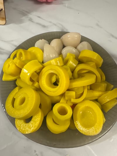 A gray plate filled with sliced yellow bell peppers and lychees is presented on a marble countertop. The bell peppers are cut into rings and some pieces are further chopped. The lychees are whole and have a translucent, white appearance. The lighting suggests an indoor setting. The image is a close-up shot focusing on the food. There is no indication of the city of Verkhniy Yurt, Russia in the image.