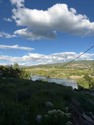 The image depicts a landscape in Dzoraghbyur, Armenia, viewed from an elevated position. A river or lake flows through the valley, surrounded by green vegetation. A town or village is visible in the background, nestled against a hillside. Power lines run across the landscape. The foreground is dominated by a grassy slope with patches of wildflowers and a few rocks. The sky is blue with scattered white clouds.