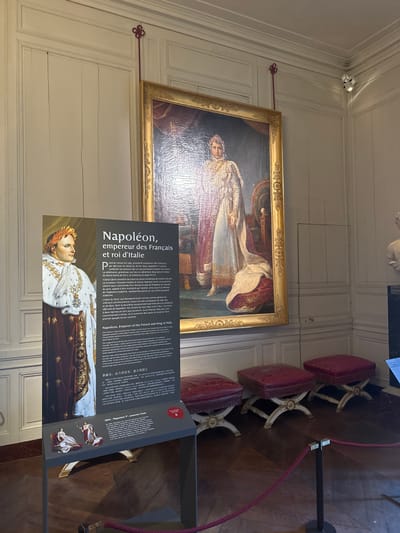 The photograph depicts an interior view in the Palace of Fontainebleau, France, where a large framed portrait of Napoleon is displayed on a white-paneled wall. The portrait is hung with an ornate golden frame. To the right of the portrait, a gray informational sign stands on the floor, bearing the title "Napoléon, empereur des Français et roi d'Italie" (Napoleon, Emperor of the French and King of Italy). The sign features an image of Napoleon and text in multiple languages, including French, English, and Chinese. To the right of the sign, three red upholstered stools with white decorative legs are visible. A red rope barrier runs along the side, preventing access. The floor is made of wood. The lighting in the room is bright.