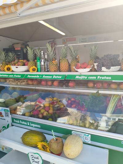 The image shows a fruit stand in Nazaré, Portugal, with a display of fresh fruits and vegetables. In the foreground, there are two melons, one green and one beige, a sunflower, and a coconut with a straw. A sign says "Fresh Fruit". The middle ground features a refrigerated display case filled with various fruits such as pineapples, peaches, grapes, apples, mangoes, dragon fruit, papayas, and watermelons, as well as some vegetables like celery and ginger. Above the display case, there are bottles and more pineapples. In the background, there is a sign that reads "Sumos Naturais" (Natural Juices) and other promotional materials. The overall scene suggests a vibrant market or shop selling healthy food options.