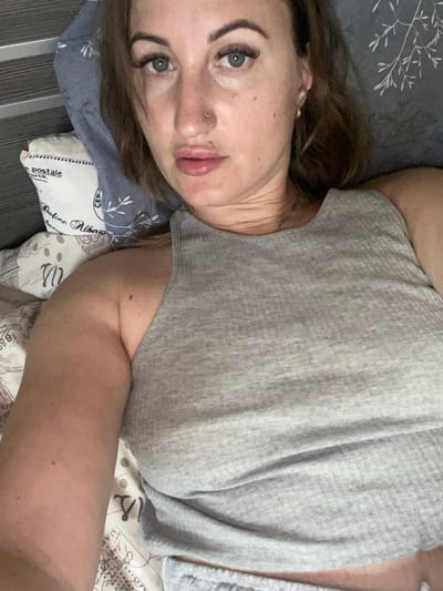 The image is a selfie taken by a woman lying in bed. She is wearing a grey tank top and has dark brown hair. The background shows a pillow with a pattern of writing and illustrations, and a grey textured wall. The woman's face is partially visible, with her eyes looking towards the camera. Her makeup includes dark eyeliner and mascara. She has a nose ring. The lighting suggests it is daytime, possibly indoors. The overall impression is a casual, personal photograph. There are no specific elements in the image that indicate a location in the city of Zarichne, Ukraine.