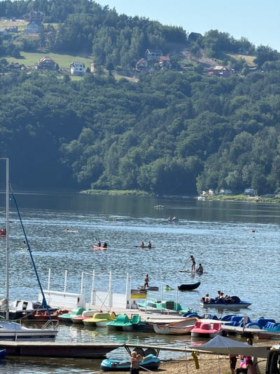 The image depicts a sunny day at a lake in Gródek nad Dunajcem, Poland. The scene is divided between a lush, tree-covered hillside with scattered houses and the glistening blue water of the lake. On the water, various individuals are engaged in recreational activities. Several people are on paddleboards, some standing, while others appear to be kayaking or paddling small boats. A cluster of colorful paddle boats are docked along a wooden pier on the right side of the image, with several people visible on and around them. The clear sky and bright sunlight suggest it is midday. The atmosphere appears relaxed and leisurely, indicative of a summer day spent enjoying outdoor water activities. The hillside is densely wooded, with several buildings nestled amongst the trees, suggesting a small village or resort area.
