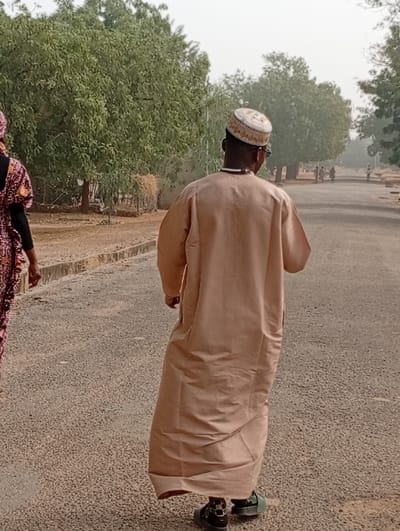 A man wearing a tan-colored robe and a small white cap walks away on an asphalt road. Trees and low vegetation are visible on both sides. Another individual in a colorful dress walks near the man. Additional people walk in the distance. The sky is overcast.
