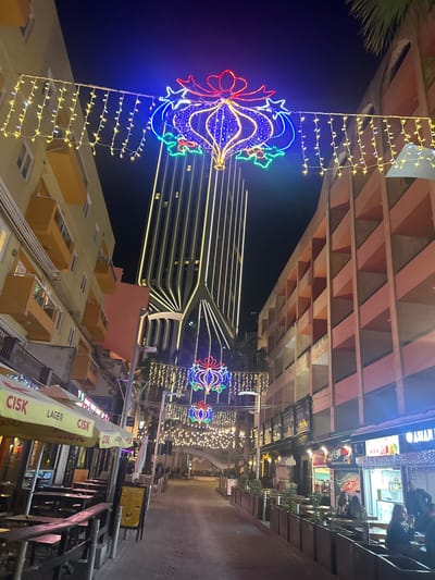 Illuminated street with Christmas ornaments. The decorations include a blue and red ornament shape with a star on top and a string of yellow lights. There is a building in the background with horizontal yellow lights. The street has shops and a shaded area with a structure above. The image is at an angle.