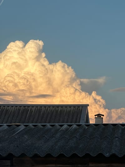 The image captures a dramatic sky scene from Primorsko-Akhtarsk, Russia, at what appears to be late afternoon or early evening, given the warm, golden light illuminating the clouds. The primary subjects are the vast, towering cumulonimbus clouds, rendered in striking shades of white and gold, hinting at their immense scale and potential for atmospheric phenomena.
A portion of a dark, corrugated roofline runs along the right side of the frame, providing a stark contrast to the ethereal clouds and grounding the scene with a sense of human presence and habitation. A small, square chimney or ventilation unit is visible atop a building in the distance, further reinforcing the impression of a residential or industrial setting. The sky itself is a clear, pale blue, creating a backdrop that emphasizes the texture and volume of the clouds.
There is no discernible activity or event taking place; the image appears to be a still moment capturing the grandeur of nature. Notable details include the powerful texture of the clouds, their illuminated edges, and the weathered appearance of the roof. The overall emotion evoked is one of awe and a sense of the sublime, juxtaposing the raw power of the weather with the mundane structures of human life. No visible text is present in the image.