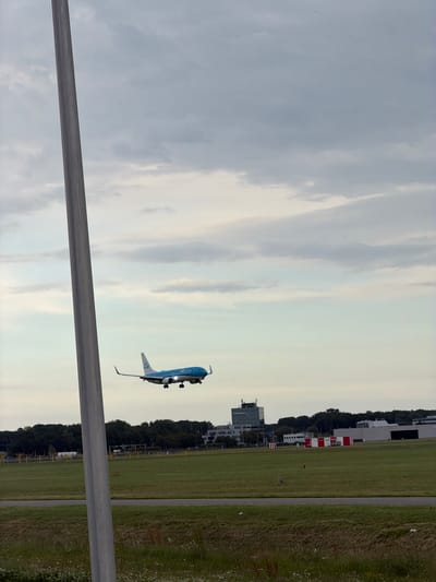 An aerial view of an airplane landing on the runway at Schiphol Airport in Schiphol, Netherlands. The airplane is a blue and white KLM jet with its landing gear down. The runway is surrounded by green grass and trees, with several buildings in the background. The sky is overcast with gray clouds. A tall, silver pole is in the foreground on the left side of the image.