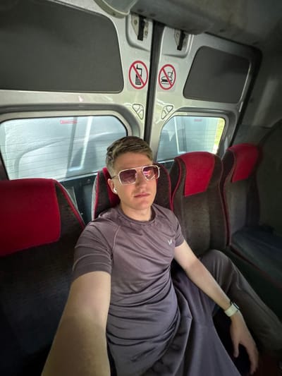 A man wearing sunglasses and a watch is reclining across three seats in what appears to be the interior of a bus or train. The seats are upholstered in gray fabric with red headrests. The interior walls are silver and feature signs indicating that certain items are prohibited. Through the windows, trees can be seen outside. The text "Notausstieg" can be read on one of the windows. The location of Staro Oriahovo, Bulgaria cannot be inferred from the image.